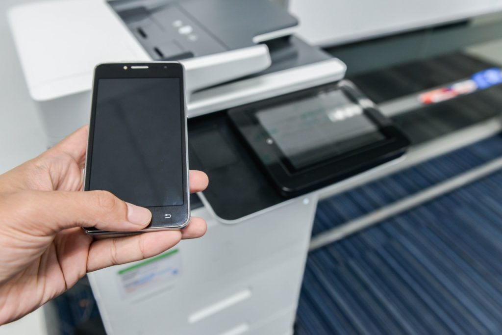 A hand holding a smartphone near a printer's control panel
